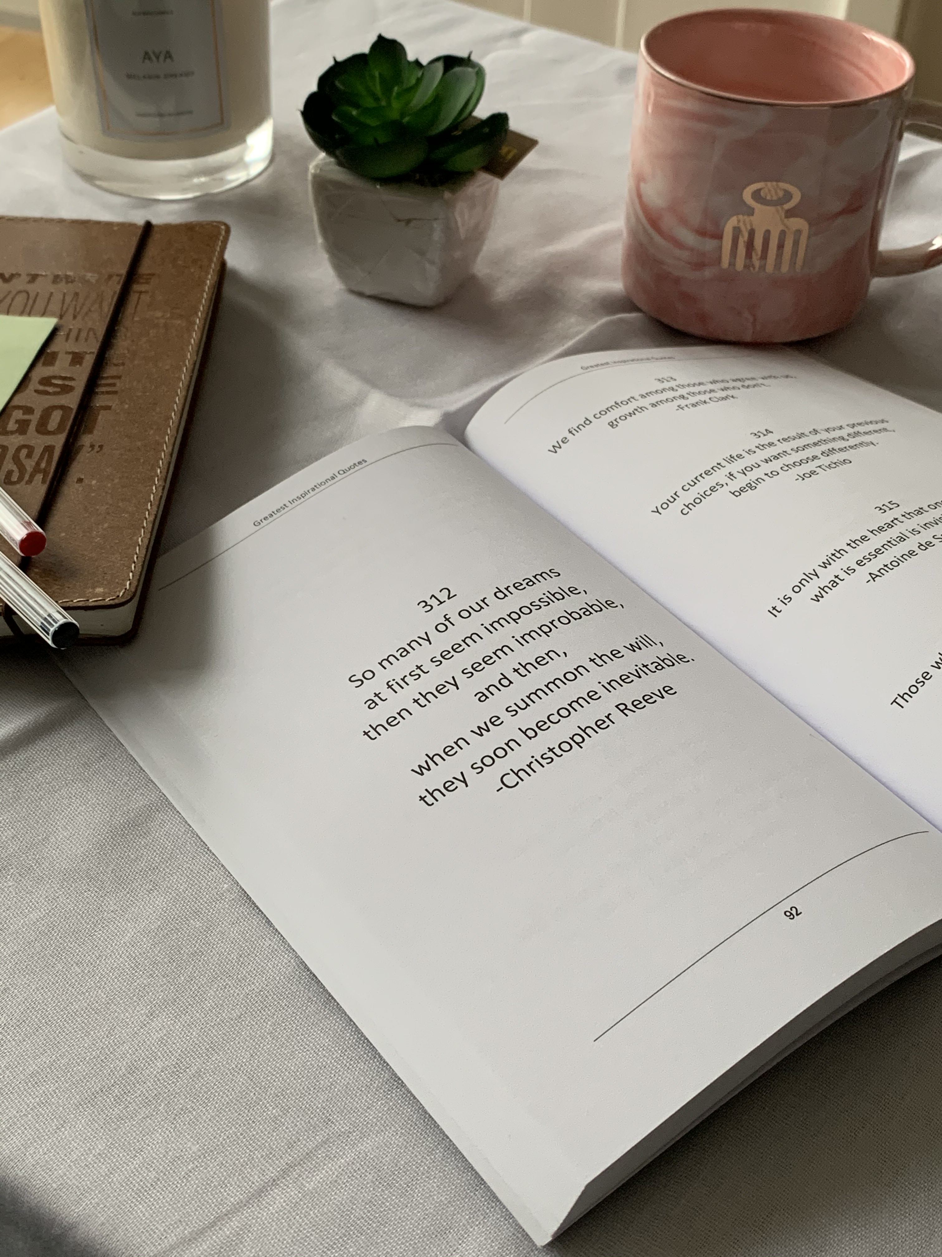 An open book with a quote with a mug, an artificial plant, and a candle in the background.